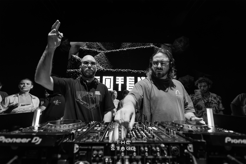 兩位DJ在聚光燈下混音，充滿活力的現場表演場景，後方有電子音樂頻道的背景。